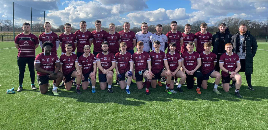 A group of males wearing burgundy shirts stand and sit around a silver trophy on grass.